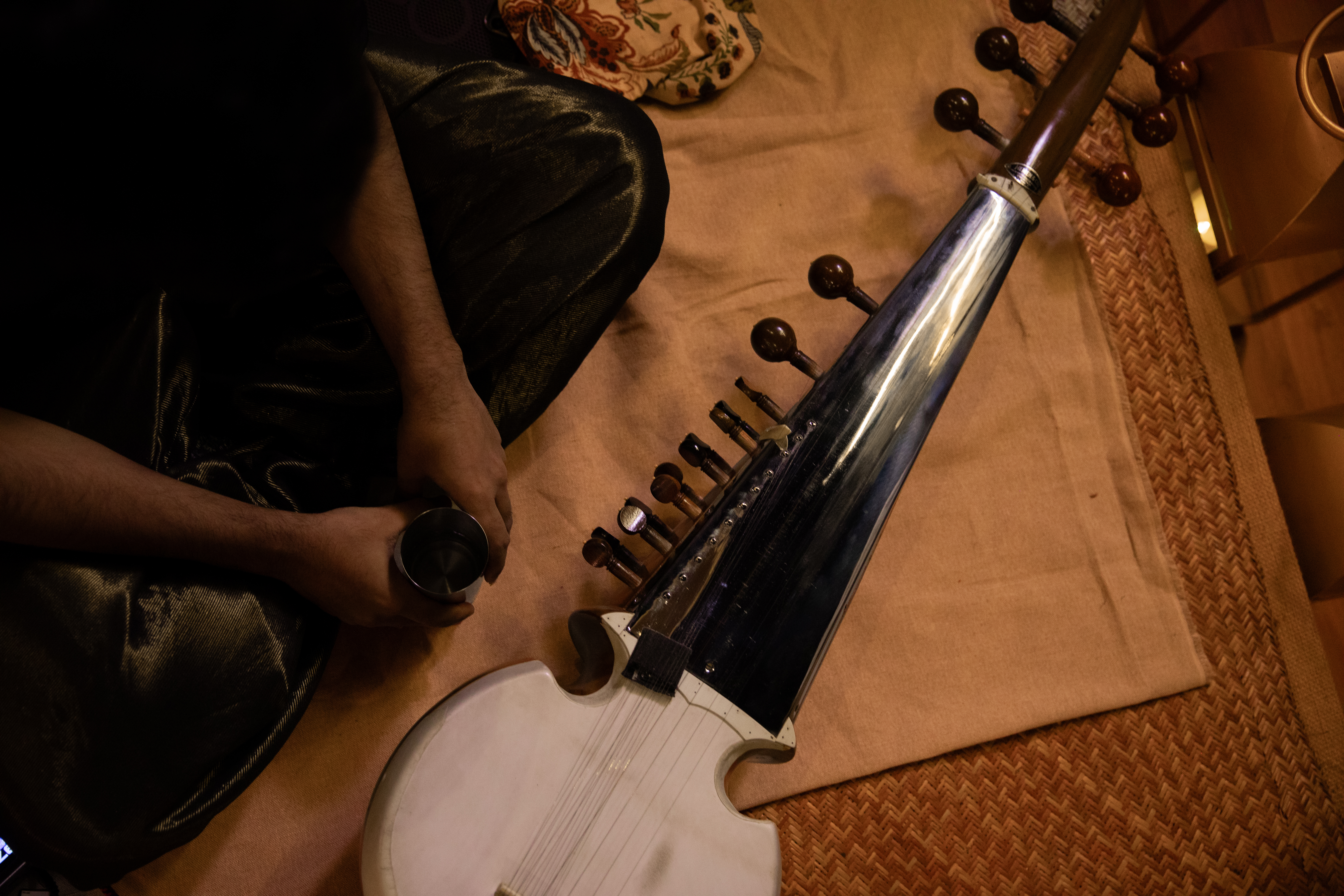 Sarod resting on woven floor mats, hands holding a metal cup, warm studio light from above.